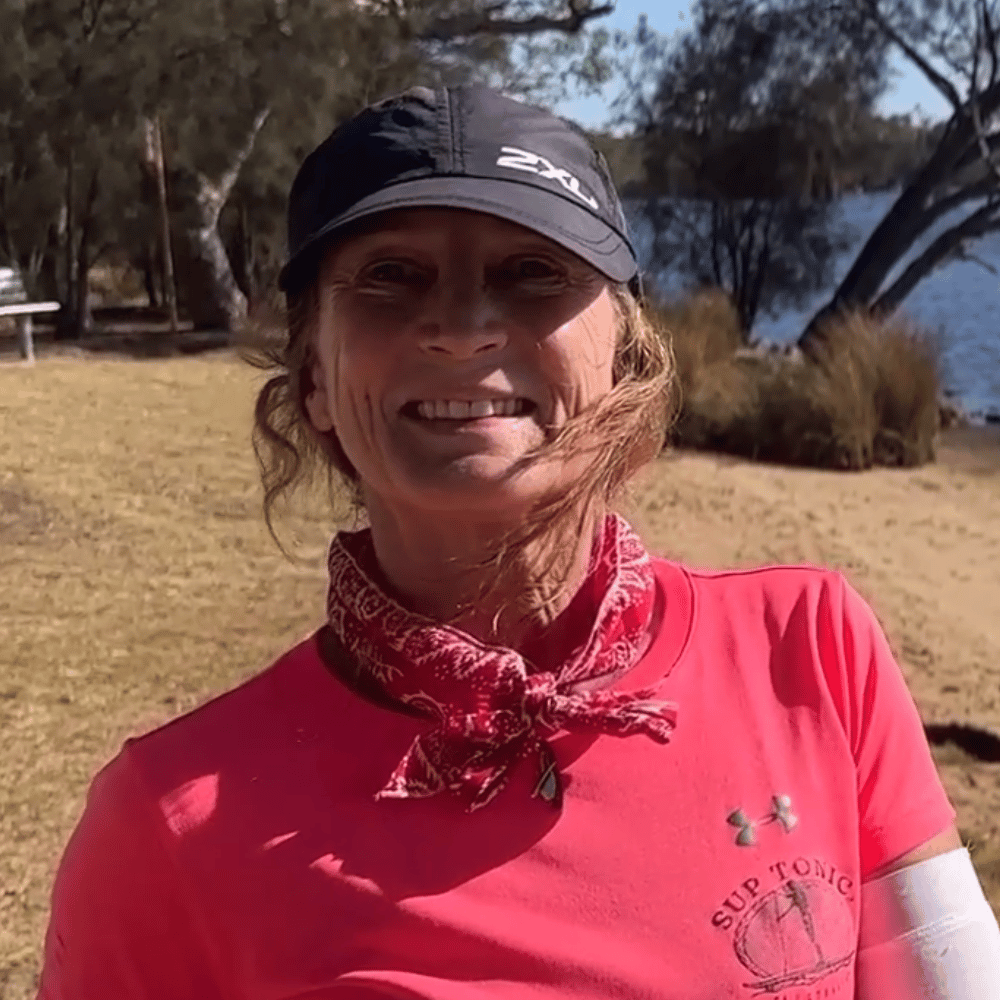 A photo of Maree wearing her SupTonic rashie and hat, standing on the banks of the Swan River in Bayswater