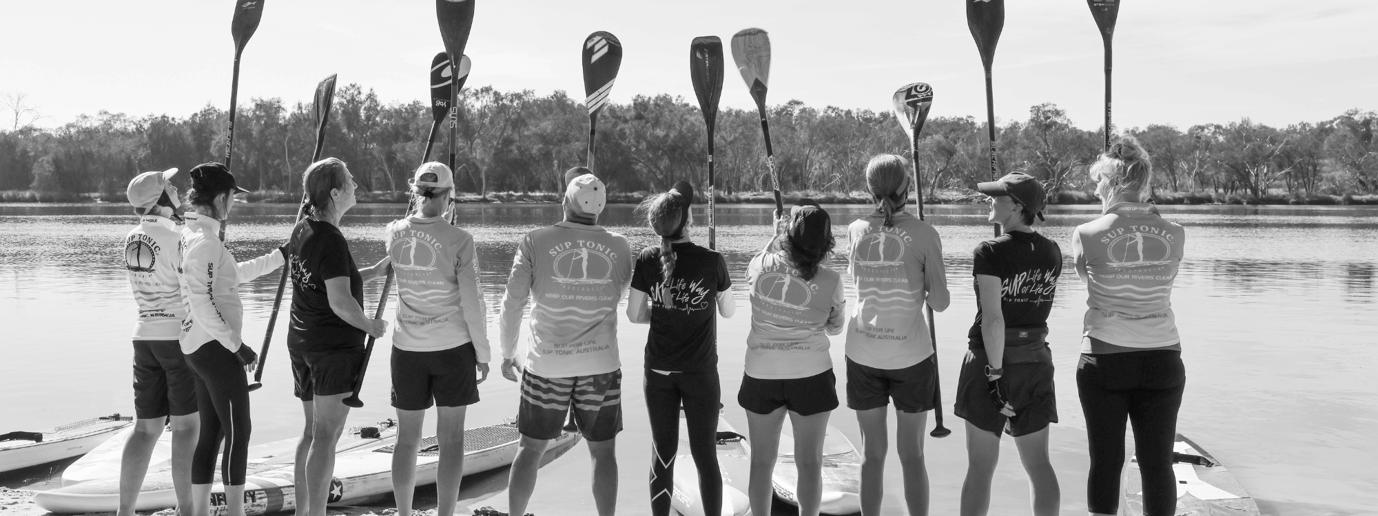 a black and white photo of a group of Stand Up Paddle Boarders on the banks of the Swan River holding thier paddles in the air, wearing SUPTONIC merchandise