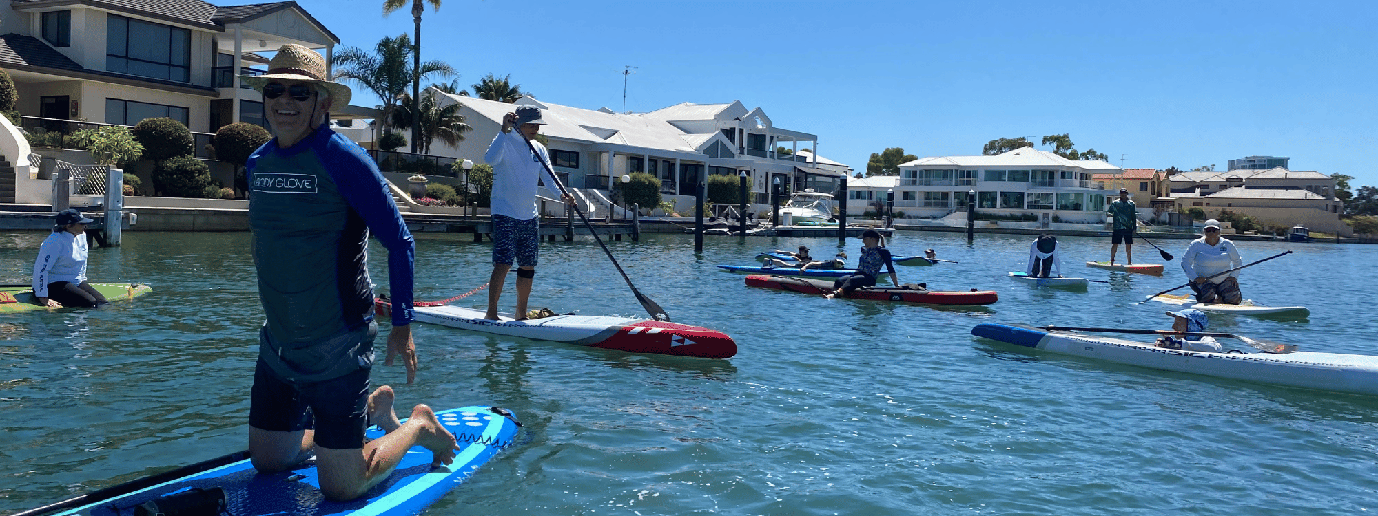 a man in a blue rashie looks very happy kneeling on a sup board, while 9 others paddle and sit on boards behind him.