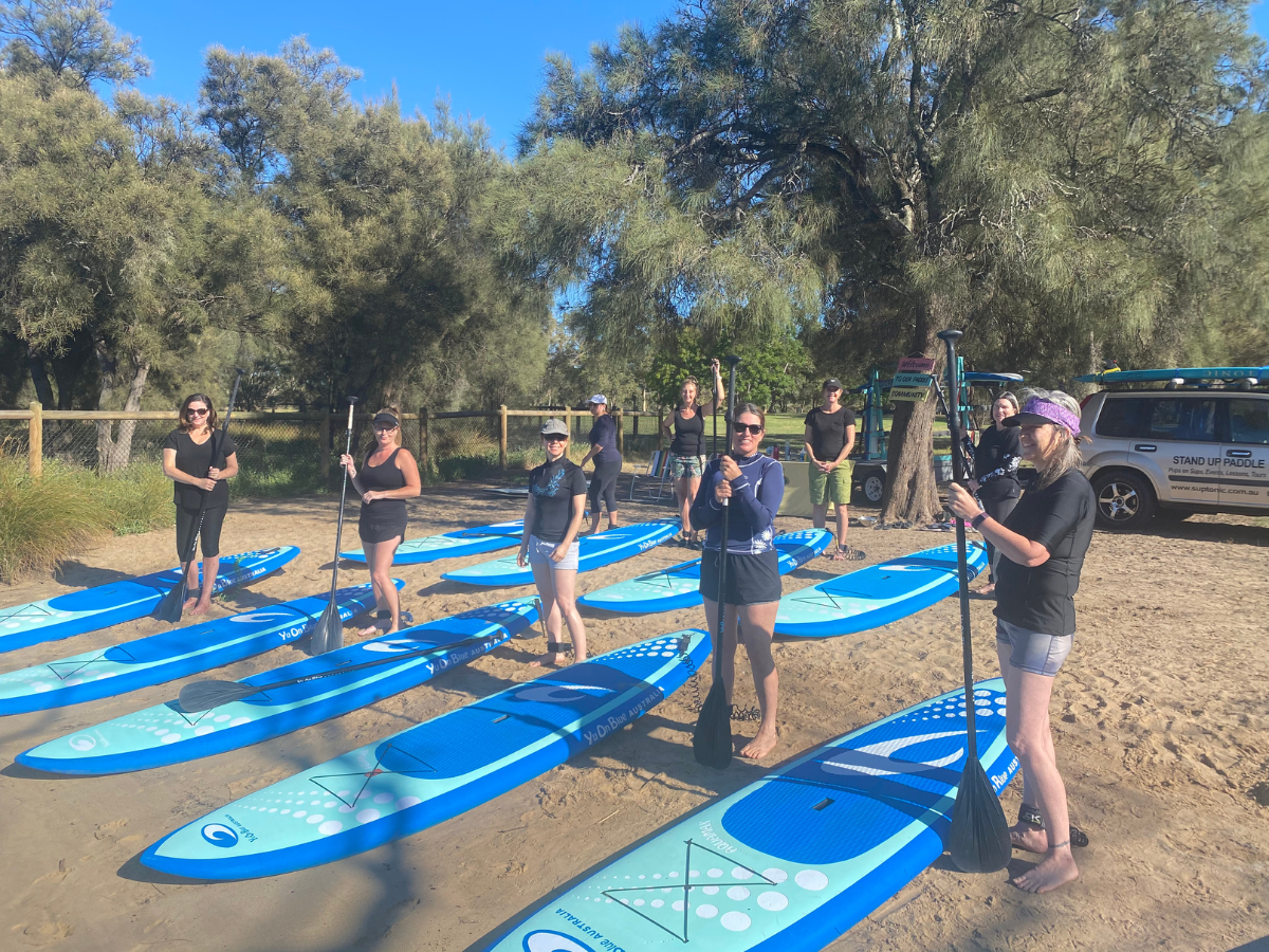 A group of new participants stand along side their blue and green paddle boards listening to instructions