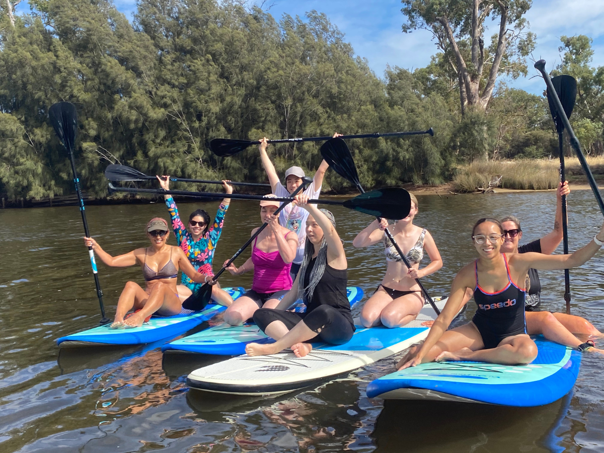 A group of women sitting and kneeling on their paddleboards all looking joyful with their paddles in the air in celebration