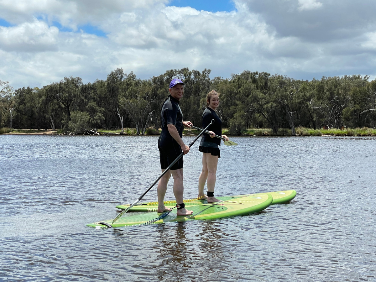 A couple standing on green stand up paddleboards looking back at the camera with big smiles on thier faces.